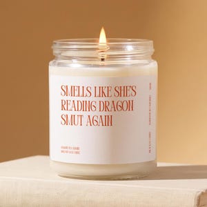 May include: A clear glass jar candle with a white label featuring the orange text "SMELLS LIKE SHE'S READING DRAGON SMUT AGAIN". The candle contains off-white wax and has a single lit wick. The label also includes the text "WHITE TEA + JASMINE" and "100% SOY WAX CANDLE" in smaller orange font.