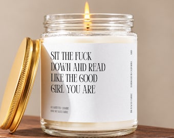 May include: A clear glass candle jar with a gold-colored metal lid, filled with white wax and a lit wick. The white label features black text that reads: SIT THE FUCK DOWN AND READ LIKE THE GOOD GIRL YOU ARE, along with smaller text details.