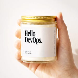 May include: A clear glass jar candle with a gold lid, held in a hand. The label reads "Hello, DevOps." and "ink n wax candle, handmade in california." The candle is a soy wax blend with Rose-Patchouli fragrance.