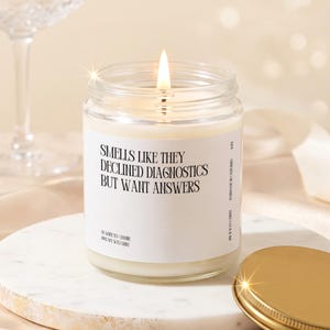 May include: A lit soy wax candle in a clear glass jar with a white label featuring the black text: SMELLS LIKE THEY DECLINED DIAGNOSTICS BUT WANT ANSWERS. The label also includes the text: OIL WHITE TEA + JASMINE, 100% SOY WAX CANDLE, HANDMADE IN CALIFORNIA.