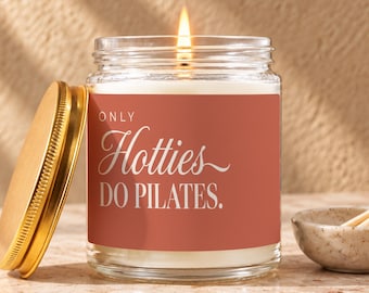 May include: A clear glass candle jar with a lit wick and a gold-colored metal lid. The label is terracotta-colored with white text that reads: ONLY Hotties DO PILATES. The candle wax is white and fills the cylindrical glass container.