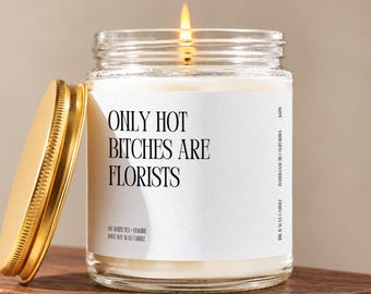 May include: A clear glass jar candle with a gold lid and a white label featuring the black text ONLY HOT BITCHES ARE FLORISTS. The candle contains white wax and has a lit wick with a visible flame, emitting a warm glow from the center of the jar.