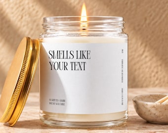 May include: A clear glass jar candle with a lit wick and a gold-colored metal lid leaning against the side. The white label features the black text "SMELLS LIKE YOUR TEXT" along with "OH WHITE TEA + JASMINE", "100% SOY WAX CANDLE", and "HANDMADE IN CALIFORNIA".
