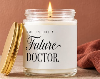 May include: A clear glass candle jar with a white label featuring the black text "SMELLS LIKE A Future DOCTOR." The candle has a lit wick and a separate gold-colored metal lid resting beside it on a textured surface.