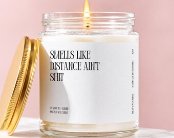 May include: A lit soy wax candle in a clear glass jar with a gold-colored metal lid. The white label features black text reading: SMELLS LIKE DISTANCE AIN'T SHIT, 03 WHITE TEA + JASMINE, 100% SOY WAX CANDLE, HANDMADE IN CALIFORNIA, and K221.