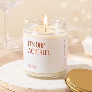 May include: A clear glass jar candle with a white label featuring the orange text IT'S DNP ACTUALLY. The candle contains white wax and has a lit wick in the center. A gold-colored metal lid is placed to the side of the candle on a round base.
