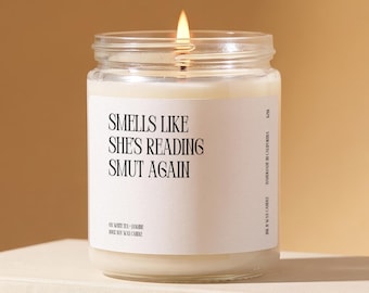 May include: A clear glass jar candle with a lit wick and white wax. The white label features black text reading: SMELLS LIKE SHE'S READING SMUT AGAIN, along with smaller text indicating white tea and jasmine scent and 100% soy wax candle composition.