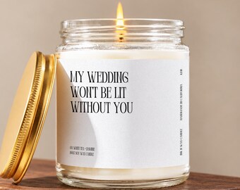 May include: A clear glass jar candle with a lit wick and a gold-colored metal lid. The white label features black text that reads: MY WEDDING WON'T BE LIT WITHOUT YOU, along with smaller text indicating soy wax and white tea and jasmine scent.