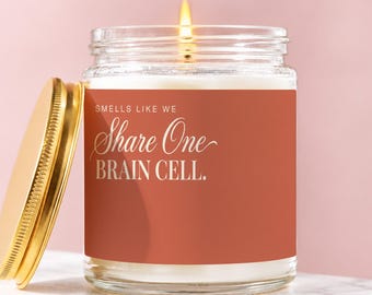 May include: A clear glass jar candle with a lit wick and a gold-colored metal lid placed to the side. The candle features a terracotta-colored label with white text that reads: SMELLS LIKE WE Share One BRAIN CELL.