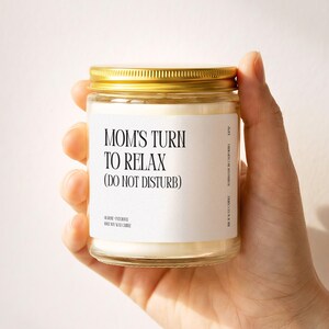 May include: A clear glass candle jar with a gold metal lid containing white wax. The white label features black text reading: MOM'S TURN TO RELAX (DO NOT DISTURB), along with smaller text indicating rose, patchouli, and 100% soy wax candle.