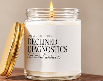 May include: A clear glass jar candle with a gold metal lid and a white label. The label features black text that reads: SMELLS LIKE THEY DECLINED DIAGNOSTICS but want answers. The candle has a single lit wick and white wax inside the glass container.