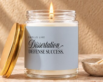 May include: A clear glass candle jar with a light blue label featuring the black text "SMELLS LIKE Dissertation DEFENSE SUCCESS." The candle has a lit wick and a gold-colored metal lid resting beside it on a light-colored stone surface.