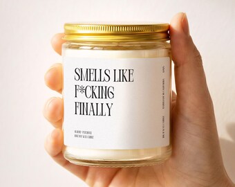 May include: A clear glass jar candle with a gold metal lid, filled with white soy wax. The white label features black text that reads: SMELLS LIKE F*CKING FINALLY, along with smaller text indicating rose, patchouli, and 100% soy wax candle.