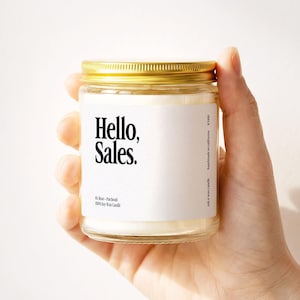 May include: A clear glass jar candle with a gold-colored metal lid. The white label features the black text "Hello, Sales." along with "01. Rose + Patchouli", "100% Soy Wax Candle", and "ink n wax candle handmade in california K1100".