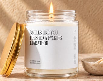 May include: A clear glass jar candle with a lit wick and a gold-colored metal lid. The white label features black text that reads: SMELLS LIKE YOU FINISHED A F*CKING MARATHON, OH WHITE TEA + INSURE, 100% SOY WAX CANDLE, HANDMADE IN CALIFORNIA.