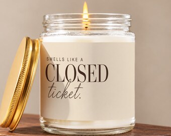 May include: A clear glass jar candle with a lit wick and a gold-colored metal lid placed to the side. The label is cream-colored with black text that reads: SMELLS LIKE A CLOSED ticket. The candle wax inside the jar is white and opaque.