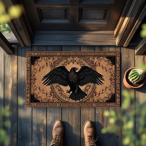 May include: Rectangular doormat featuring a black raven with outstretched wings, centered on a tan background with ornate, dark brown border. The doormat is placed on a wooden porch, with a small potted cactus visible.