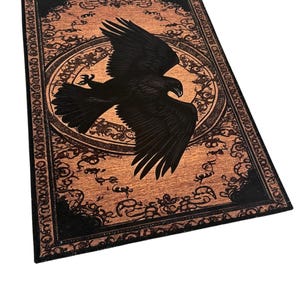 May include: A rectangular, decorative rug featuring a black eagle in flight against a tan background. The rug has a black border with ornate, swirling patterns. A circular design surrounds the eagle, adding to the rug's detailed aesthetic.