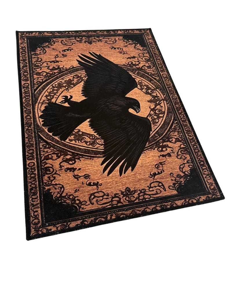 May include: A rectangular, decorative rug featuring a black eagle in flight against a tan background. The rug has a black border with ornate, swirling patterns. A circular design surrounds the eagle, adding to the rug's detailed aesthetic.