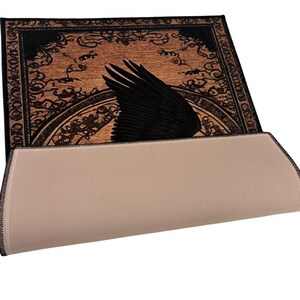 May include: A rectangular rug with a black raven wing design on a brown background. The wing is prominently displayed against a backdrop of ornate, swirling patterns. The rug's underside is a light beige color.
