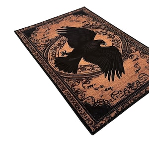 May include: Rectangular rug with a detailed black eagle design on a copper-colored background. The eagle is centered within a circular frame, surrounded by ornate patterns. The border is black, adding contrast to the overall design.