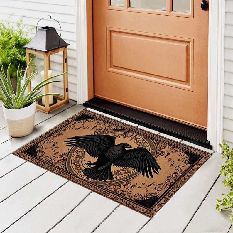 May include: A rectangular doormat featuring a detailed black raven with outstretched wings, centered on a tan background. The raven is surrounded by a circular design and ornate black border. The mat is placed in front of a brown door.