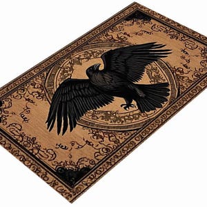 May include: A rectangular, tan-colored decorative mat featuring a detailed illustration of a black raven with outstretched wings. The raven is centered within a circular design, surrounded by ornate, dark brown patterns and text. The border of the mat is also decorated with similar patterns.