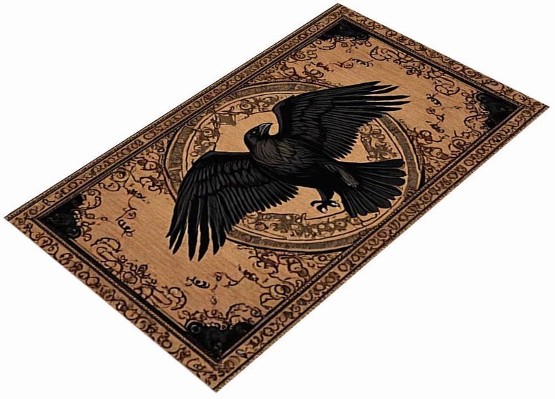 May include: A rectangular, tan-colored decorative mat featuring a detailed illustration of a black raven with outstretched wings. The raven is centered within a circular design, surrounded by ornate, dark brown patterns and text. The border of the mat is also decorated with similar patterns.