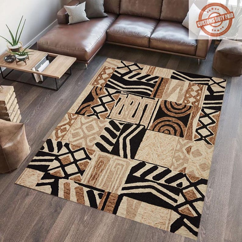 May include: A rectangular area rug with a patchwork design. The rug features geometric patterns in black, tan, and brown. The rug is placed on a hardwood floor, with a brown leather sectional sofa and a wooden coffee table in the background.