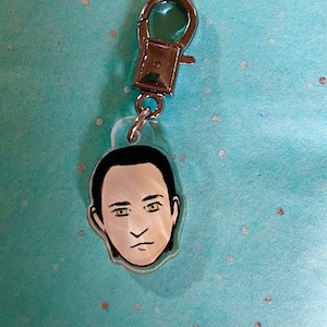 Könnte beinhalten: Ein silberner Schlüsselanhänger mit einem durchsichtigen Plastik-Anhänger, der eine Comic-Illustration eines Männergesichts zeigt.
