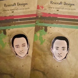 Könnte beinhalten: Zwei transparente Acryl-Pins mit einer Cartoon-Illustration eines Gesichts. Die Pins befinden sich auf einem braunen Hintergrund mit dem Text "Rexcraft Designs" und "www.roxcraft.etsy.com".