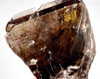 May include: A close-up of a raw, natural mineral specimen. The crystal is a deep brown color with areas of translucence, revealing internal structures and iridescent flashes of color. The surface has a rough, textured appearance.