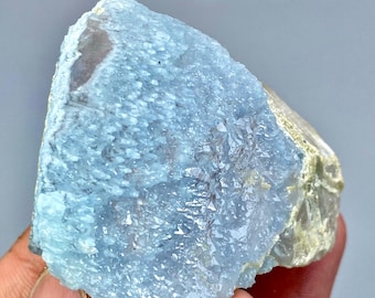May include: A rough, light blue mineral specimen with a crystalline structure. The stone has a textured surface and a mix of light blue and off-white colors. The mineral is held in a hand.