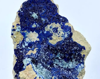 May include: A natural azurite specimen with a deep blue crystalline structure. The azurite is set against a beige matrix, creating a striking contrast. The mineral sample is irregularly shaped and displays a rough texture.