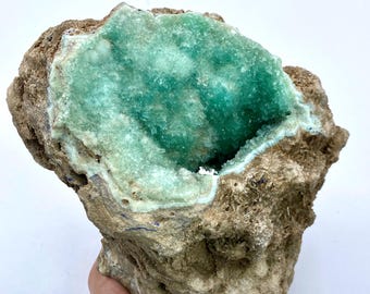 May include: A large, rough-textured rock formation with a central cavity filled with sparkling, vibrant teal crystals. The outer rock is a mix of tan and brown hues, creating a striking contrast with the crystal interior. A natural mineral specimen.