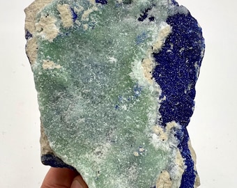 May include: A natural mineral specimen featuring a mix of colors. The stone has a central area of pale green, surrounded by dark blue and off-white sections. The mineral's surface has a textured appearance.