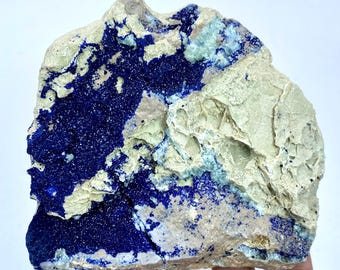 May include: A raw azurite specimen with a deep blue color, contrasted by a light beige matrix. The mineral displays a rough, textured surface with areas of crystalline formations. The stone is irregularly shaped and appears natural.