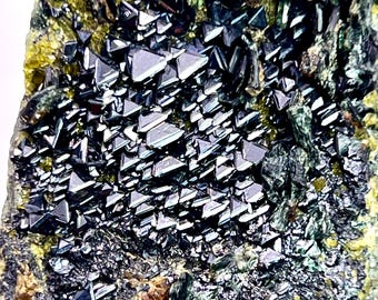 May include: Close-up of a mineral specimen featuring a cluster of lustrous, dark, triangular crystals. The crystals are set against a backdrop of green, yellow, and gray rock, creating a striking contrast. The overall texture is rough and uneven.