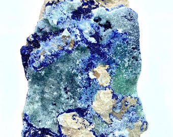 May include: A natural azurite mineral specimen with a mix of deep blue, light blue, and green hues. The stone has a rough, textured surface with patches of beige and white. The mineral is held in a hand against a white background.