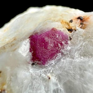 May include: A raw, deep magenta-coloured crystal embedded within a translucent, milky white mineral matrix. The central gemstone features a textured, crystalline surface, while the surrounding white stone displays natural fractures and dark inclusions.