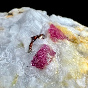 Puede incluir: Un espécimen mineral en bruto que presenta dos prominentes formaciones cristalinas de color rosa magenta vibrante incrustadas en una matriz cristalina texturizada de color blanco translúcido y amarillo pálido, junto a una pequeña inclusión metálica oscura.