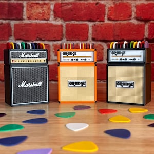 May include: Three miniature guitar amplifier replicas, one black with "Marshall" text, and two orange with "Orange" text. Colorful guitar picks are scattered on the wooden surface in front of the amps. The background is a brick wall.