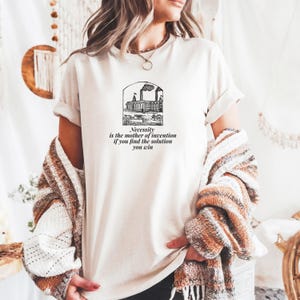 Puede incluir: Una camiseta de manga corta color crema con un gráfico negro de una fábrica con chimeneas y el texto: Necessity is the mother of invention if you find the solution you win. La camiseta tiene cuello redondo y mangas remangadas.