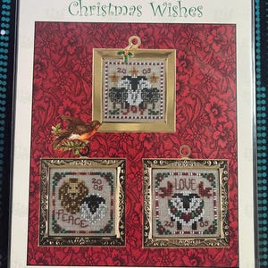 May include: A Just Nan Christmas Wishes cross stitch pattern package featuring three square framed designs with sheep, a lion, and birds. The embroidery includes the text 20 05, JOY, PEACE, and LOVE, set against a red floral background.