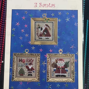 Puede incluir: Un kit de punto de cruz Just Nan 3 Santas con tres diseños cuadrados enmarcados de Santa Claus. El empaque muestra el título, el sitio web e imágenes de los proyectos de costura terminados con marcos dorados sobre un fondo de estrellas azules.