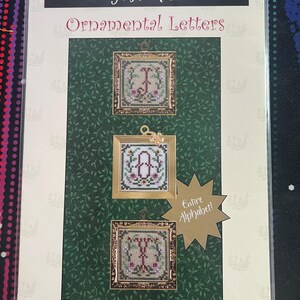 Puede incluir: Un paquete de patrones de punto de cruz Just Nan Ornamental Letters con tres diseños cuadrados enmarcados con motivos florales y letras. El empaque incluye un gráfico de estrella dorada con el texto Entire Alphabet! y un fondo verde oscuro.