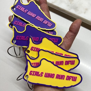 Puede incluir: Cuatro llaveros en forma de zapato amarillos y morados con el texto "Girls Who Run DFW" impreso en rosa. Los llaveros están unidos a llaveros plateados.