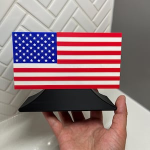 May include: A rectangular American flag with red and white stripes and a blue field of white stars. The flag is mounted on a black, angled base.