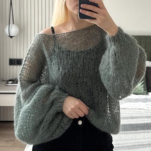 May include: A sheer, sage green knit sweater with long, voluminous sleeves. The open weave design reveals the underlayer. The sweater has a boat neck and is paired with black pants. The style is casual and comfortable.
