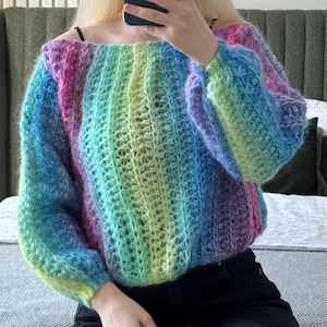 Puede incluir: Un suéter multicolor con hombros descubiertos. El suéter presenta un degradado de colores, incluyendo rosa, morado, azul, verde y amarillo. Las mangas son largas y abullonadas. El suéter está hecho de un hilo suave y texturizado.